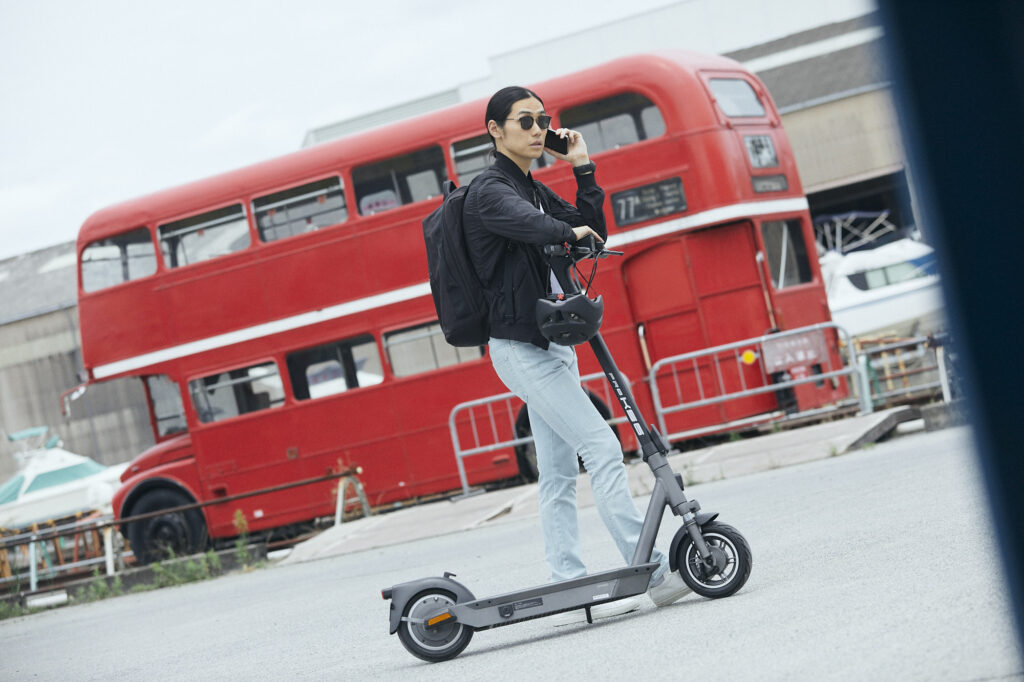 赤い二階建てバスの前で電動キックボードに乗る男性が電話している様子